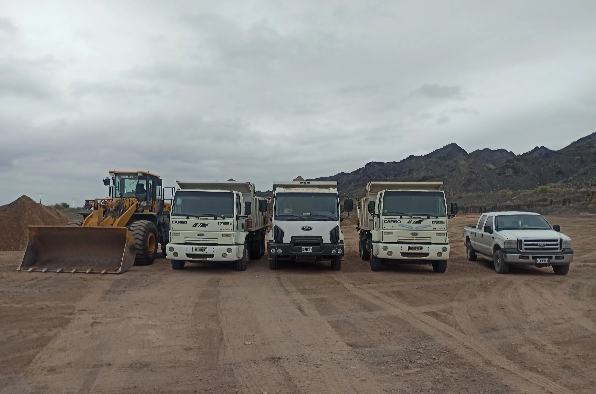 Pala cargadora, tres camiones volcadores y una camioneta alineados frente a los cerros